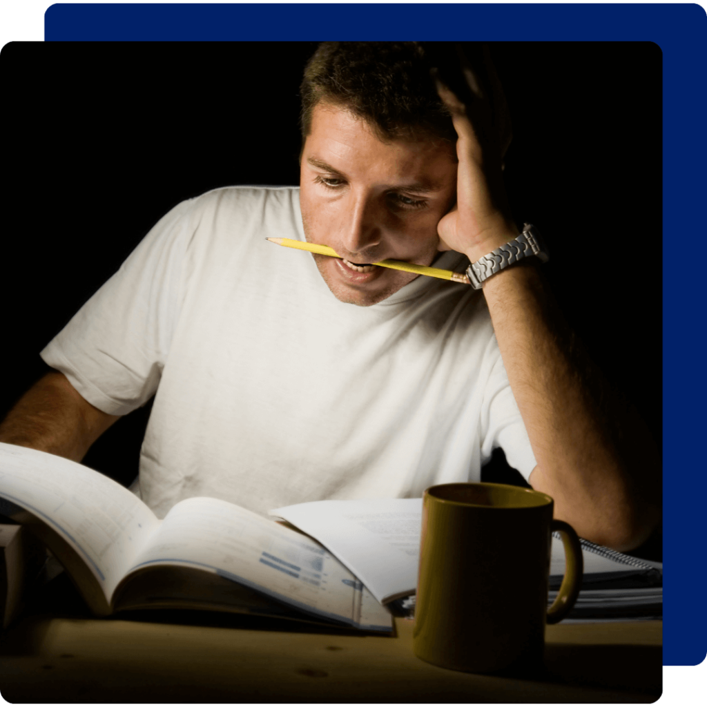 Um homem está com o lápis na boca, um livro e caderno aberto, à sua frente tem uma caneca. Sua expressão facial retrata alguém que está tendo dificuldades nos estudos e sente-se frustrado.